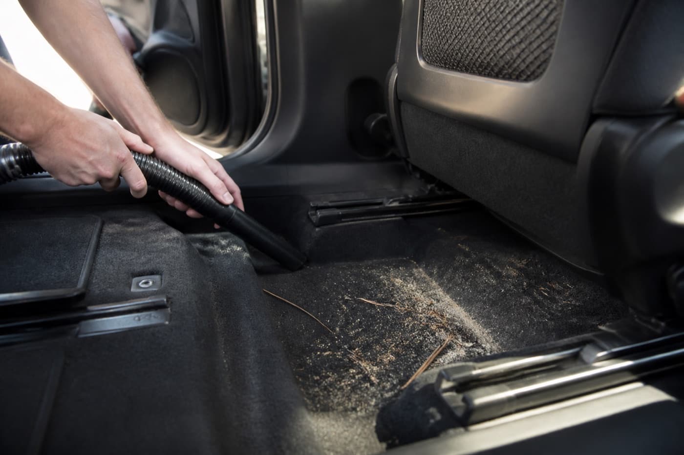 Detailer vacuuming beach sand and pine needles from a vehicle's rear footwell after a summer road trip